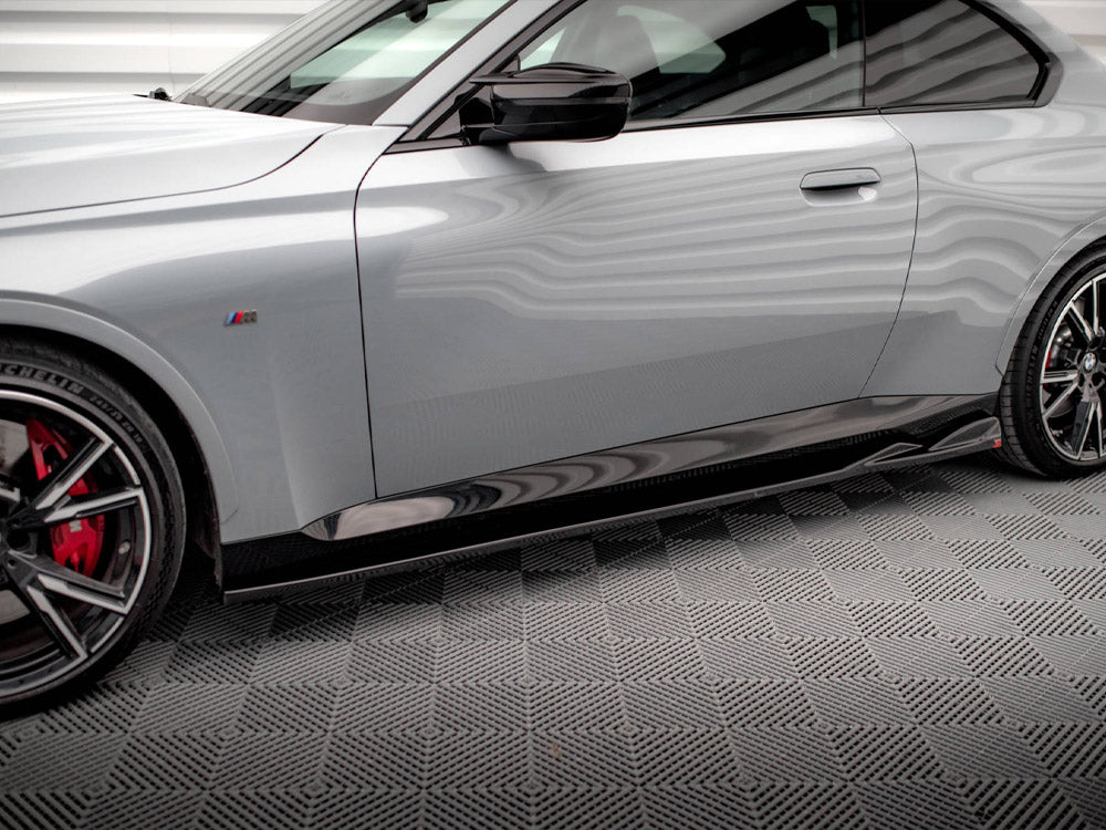 A sleek, silver car is parked on a textured gray floor. It features red brake calipers, sporty black wheels, and a side emblem with colored stripes.