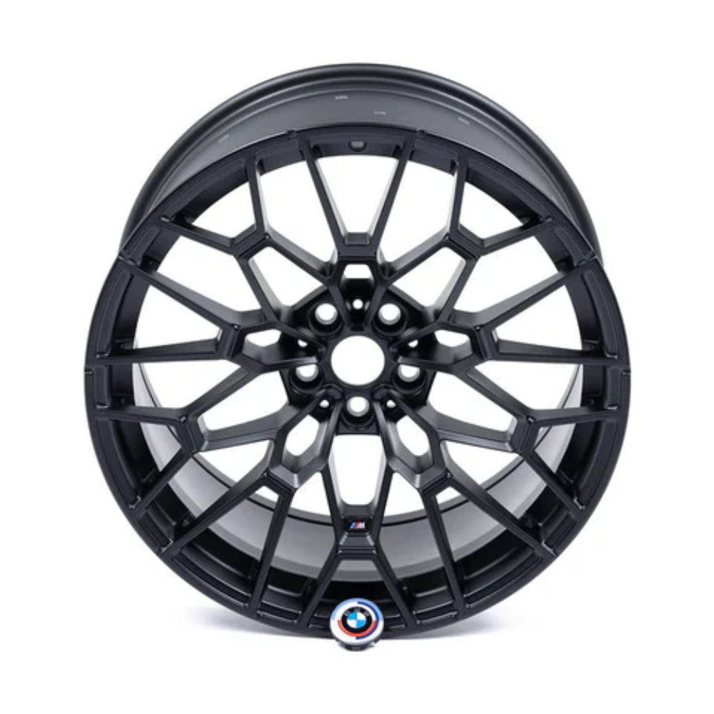 A black, intricately designed car wheel rim, featuring a multi-spoke pattern, is positioned in isolation on a white background. Near the bottom, a circular emblem displays a red and blue "M".
