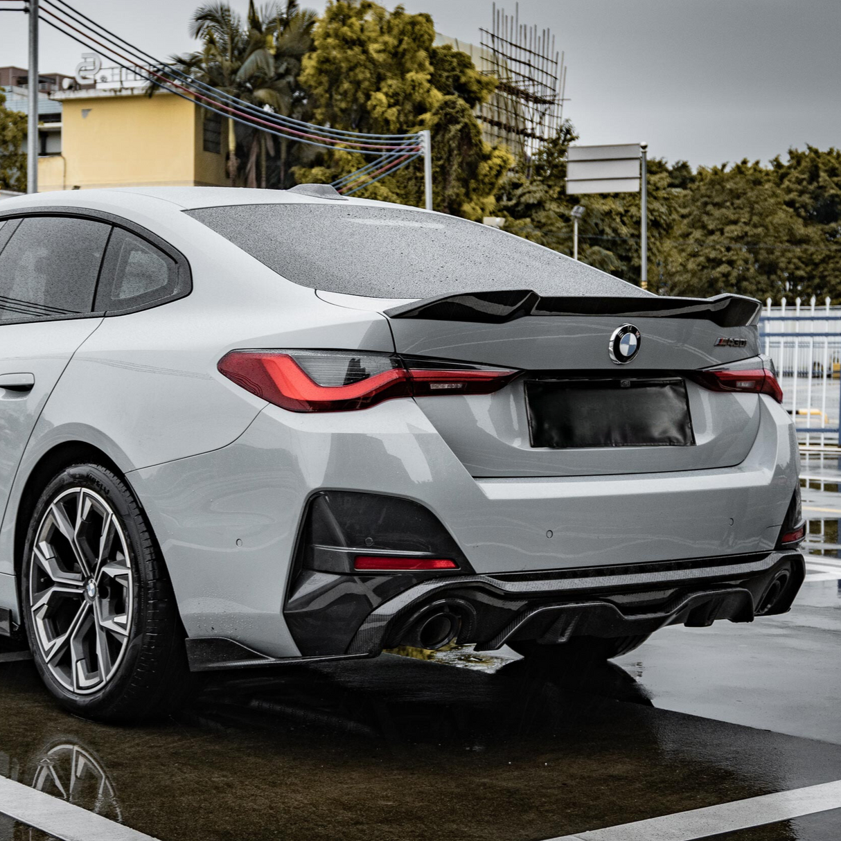 A grey sports car is parked on a wet surface, viewed from the rear, with a visible BMW logo. The background includes trees, a yellow building, and overcast sky.