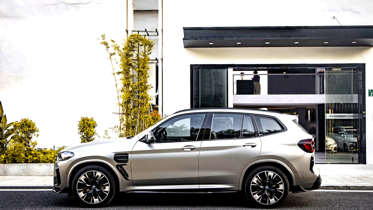 A silver SUV is parked beside a modern building with a black entrance. Yellow-green plants line the sidewalk. Reflections show other vehicles and signage: "INSTITUTO DE DRY CARBON".