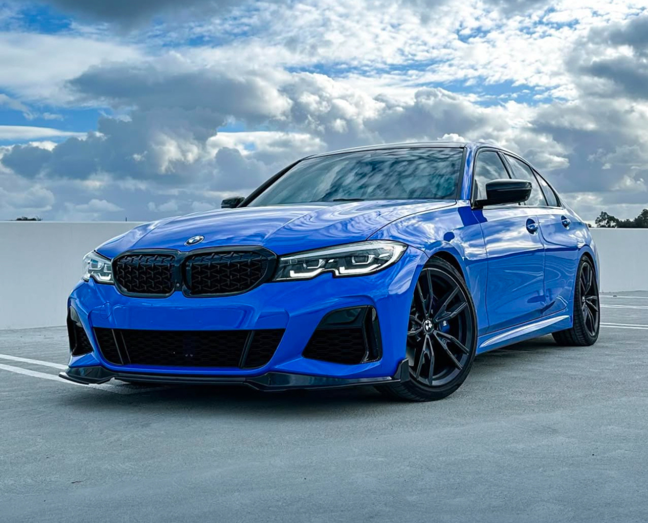 A blue BMW car is parked on a rooftop parking lot, under a cloudy sky with patches of blue.