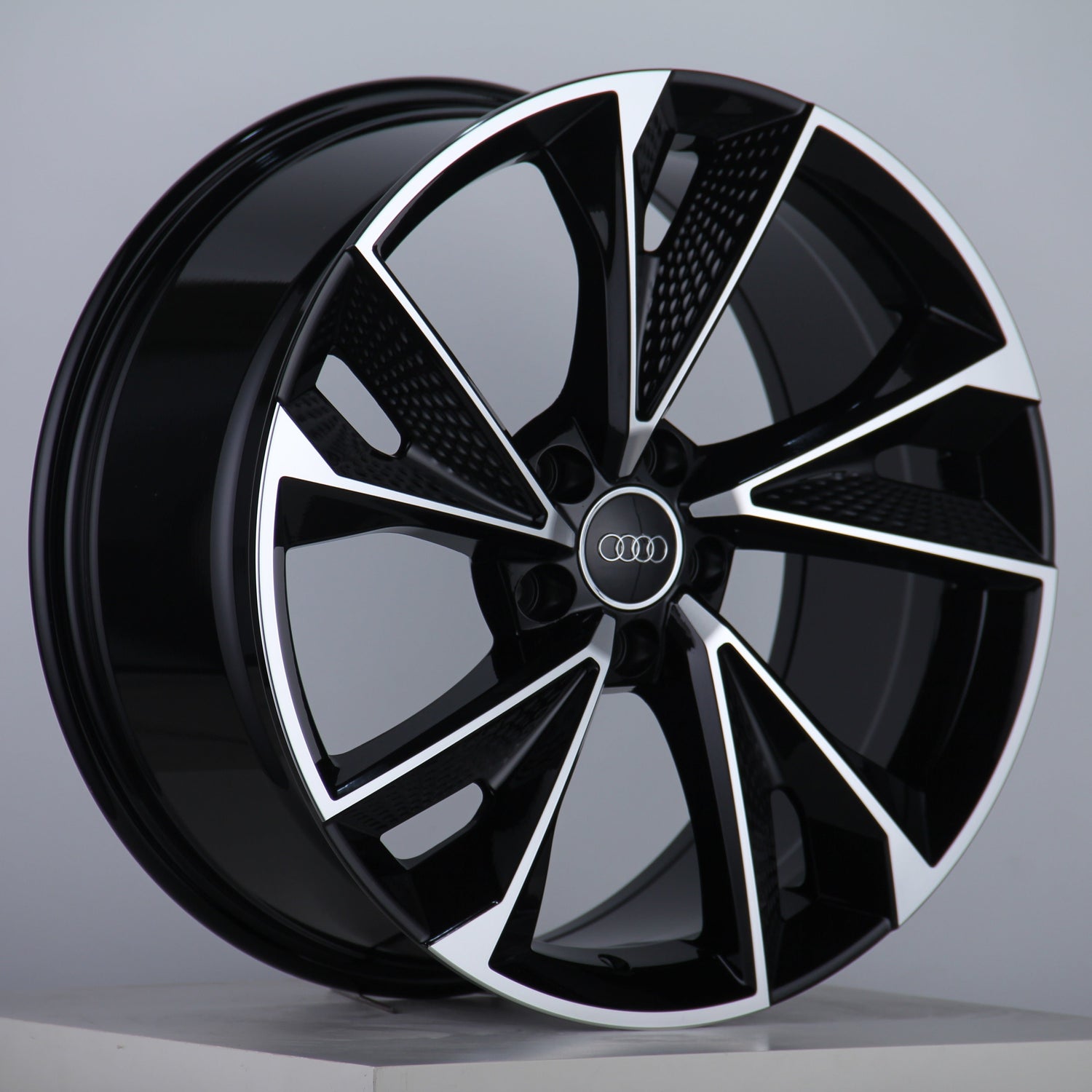 A black and silver alloy wheel with a multi-spoke design, displaying the Audi logo at the center, is positioned upright on a plain surface against a neutral background.