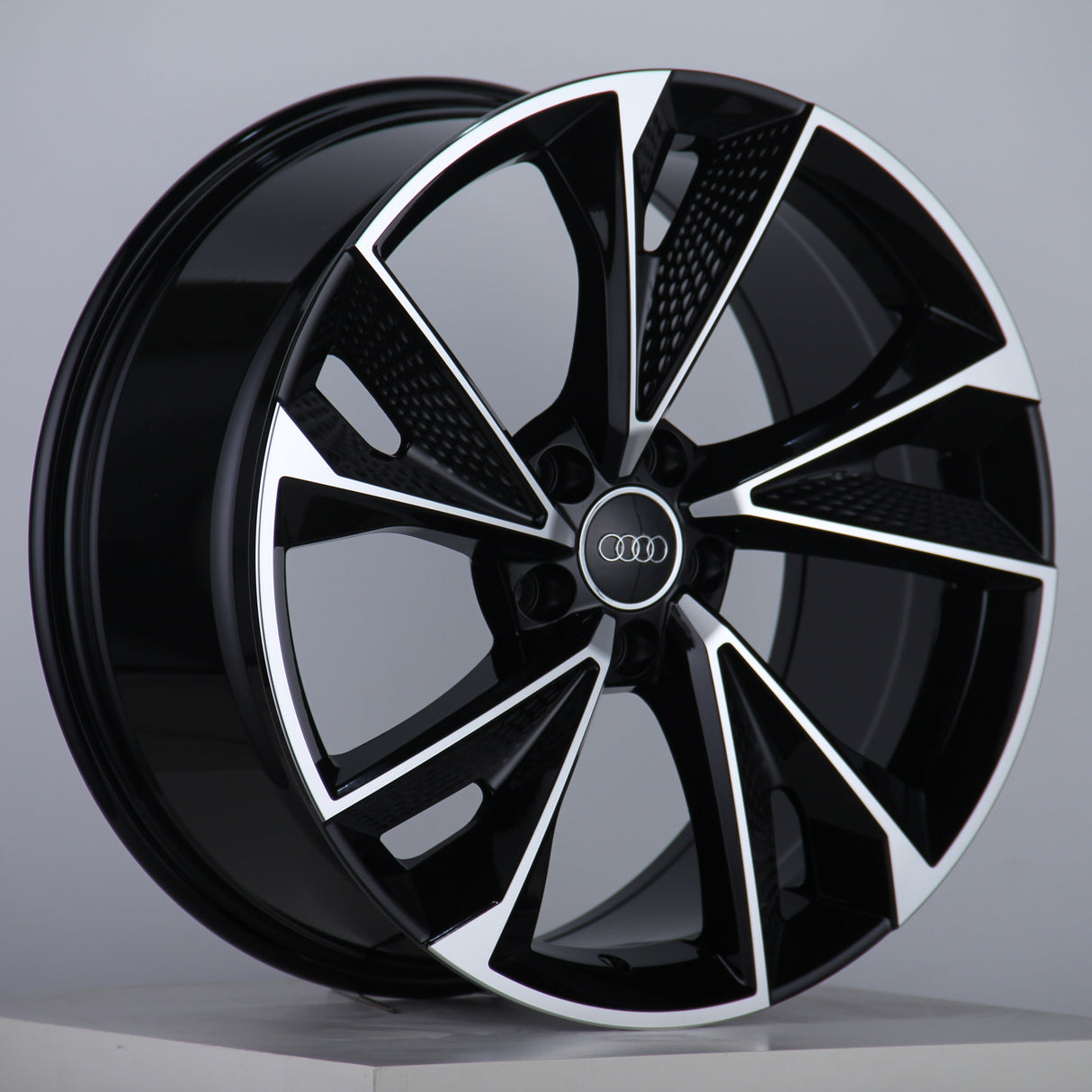 A sleek, black and silver car wheel rim with a five-spoke design, featuring the Audi logo in the center, is displayed against a plain gray background.