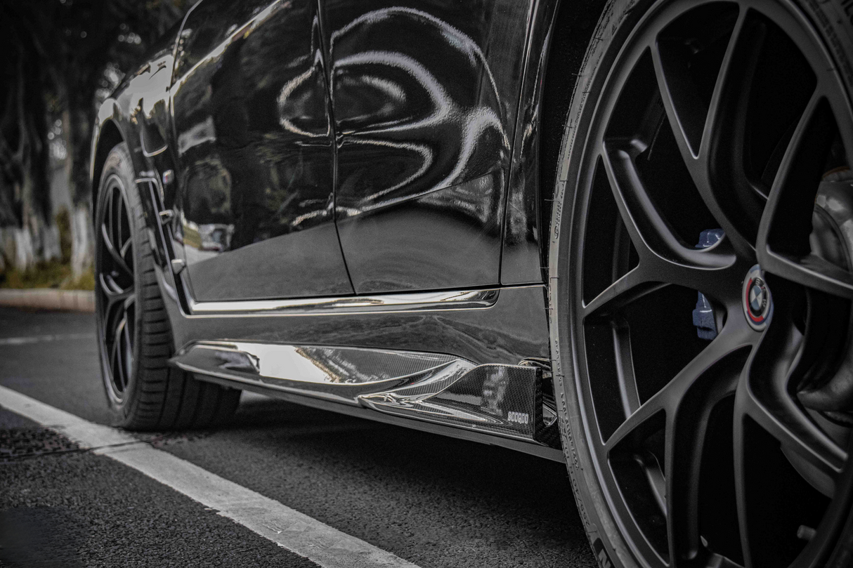 A black car's side view, showing glossy bodywork and a detailed wheel with a BMW logo, parked on asphalt near trees. Visible text includes "SOCODA."