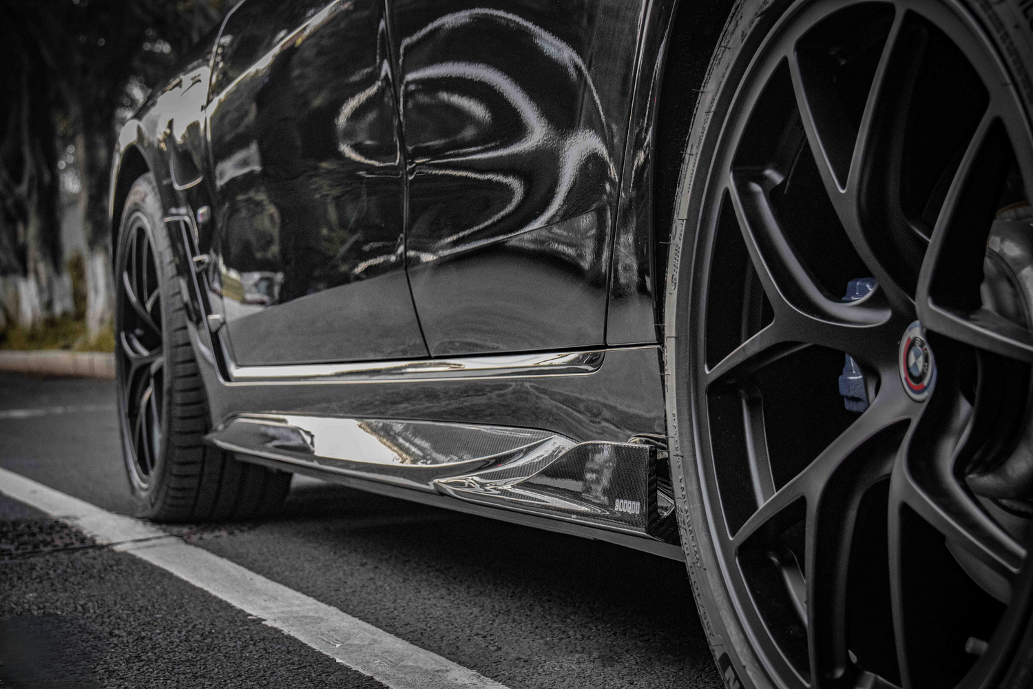 A black car's side view, showing glossy bodywork and a detailed wheel with a BMW logo, parked on asphalt near trees. Visible text includes "SOCODA."