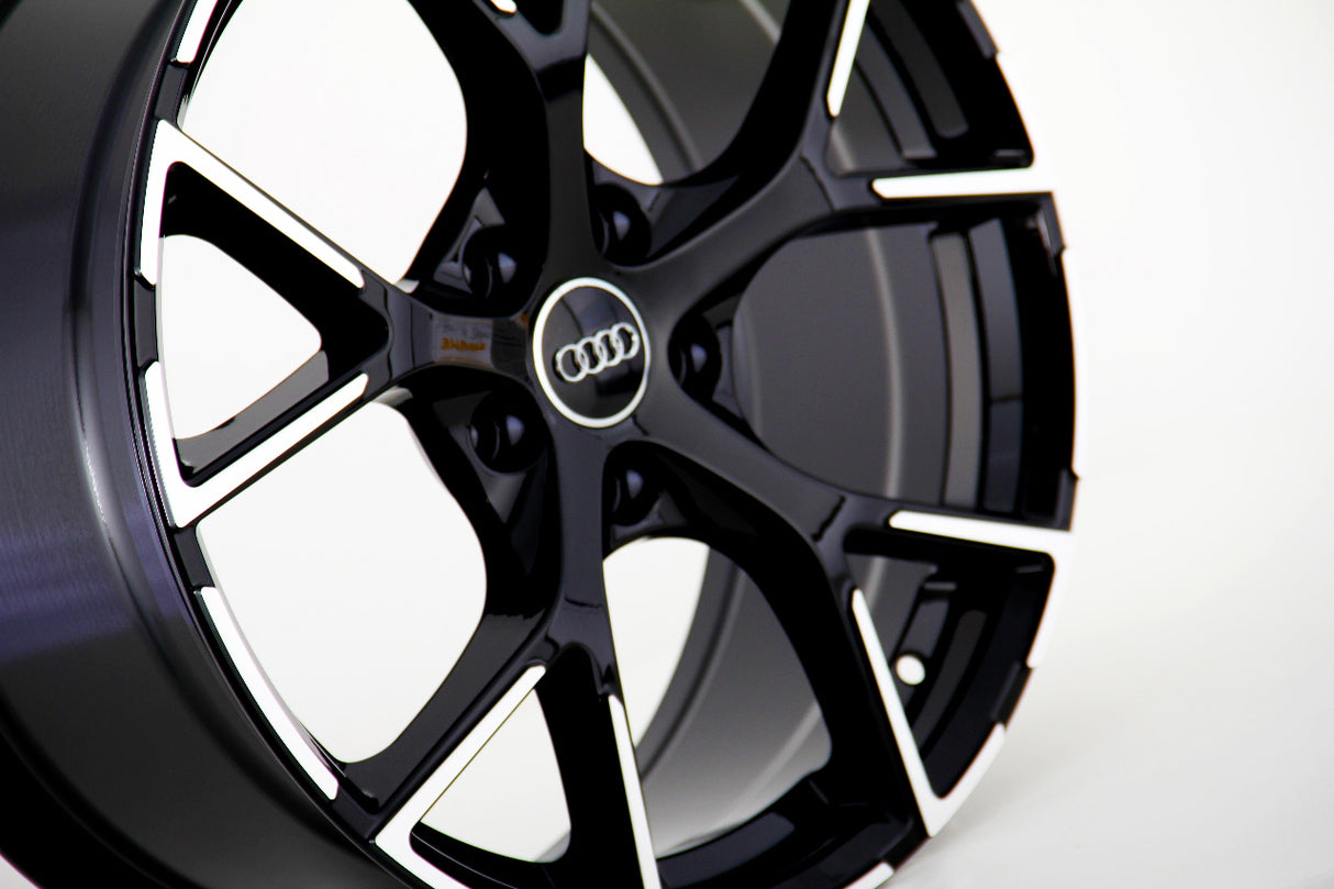 A glossy black car wheel with intricate spoke design displays an Audi logo in the center, set against a plain white background.