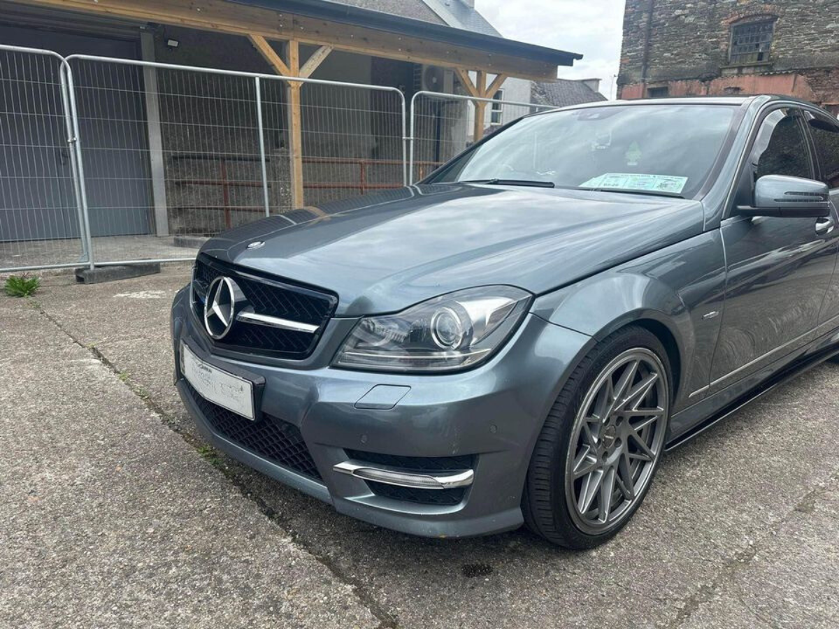 A silver Mercedes car is parked on a concrete surface near a metal fence and wooden structure. The car's windshield displays documents. The setting is an outdoor area adjacent to buildings.