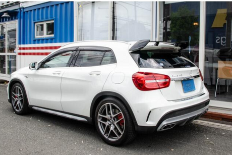 White car parked beside a blue shipping container building, displaying shiny rims and tinted windows. The background includes glass doors and a building interior with chairs.