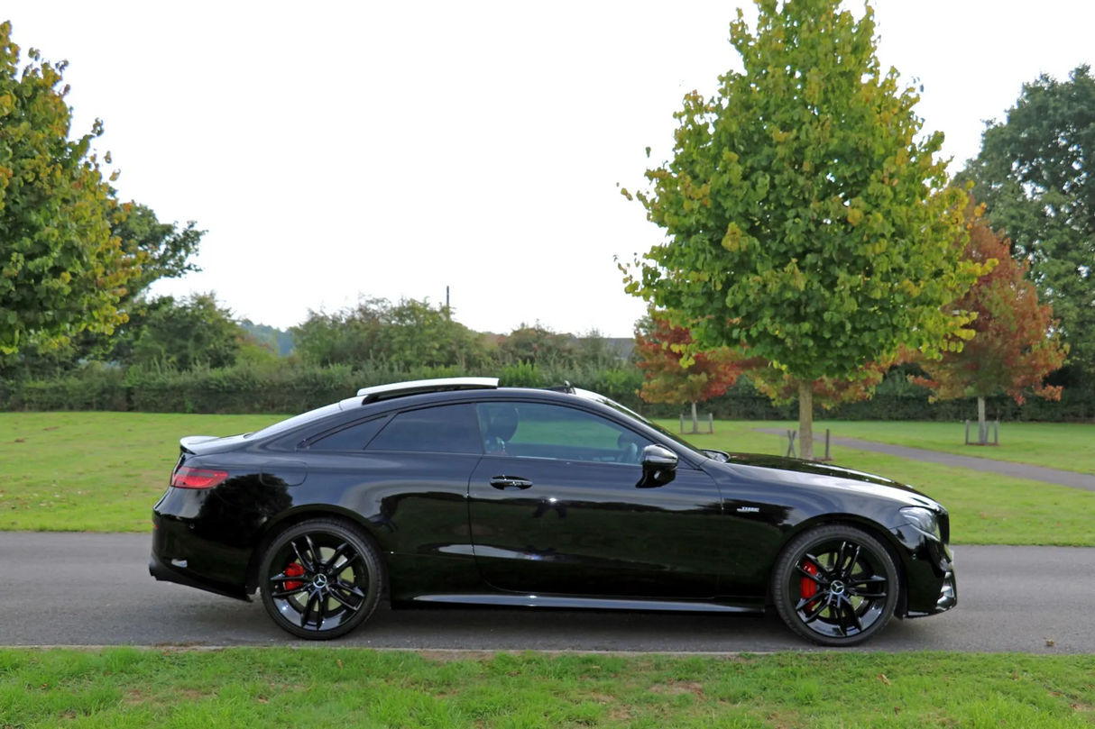 A black sports car is parked on a paved road, set against a grassy park landscape with trees in varying stages of seasonal color change.