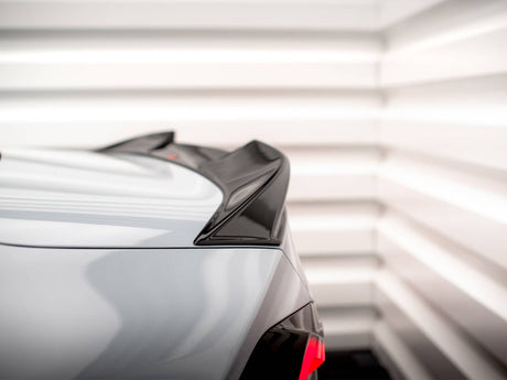 A car's rear spoiler is visible in close-up, featuring a glossy black finish. It is situated in a brightly lit, white-paneled room, enhancing its sleek design.