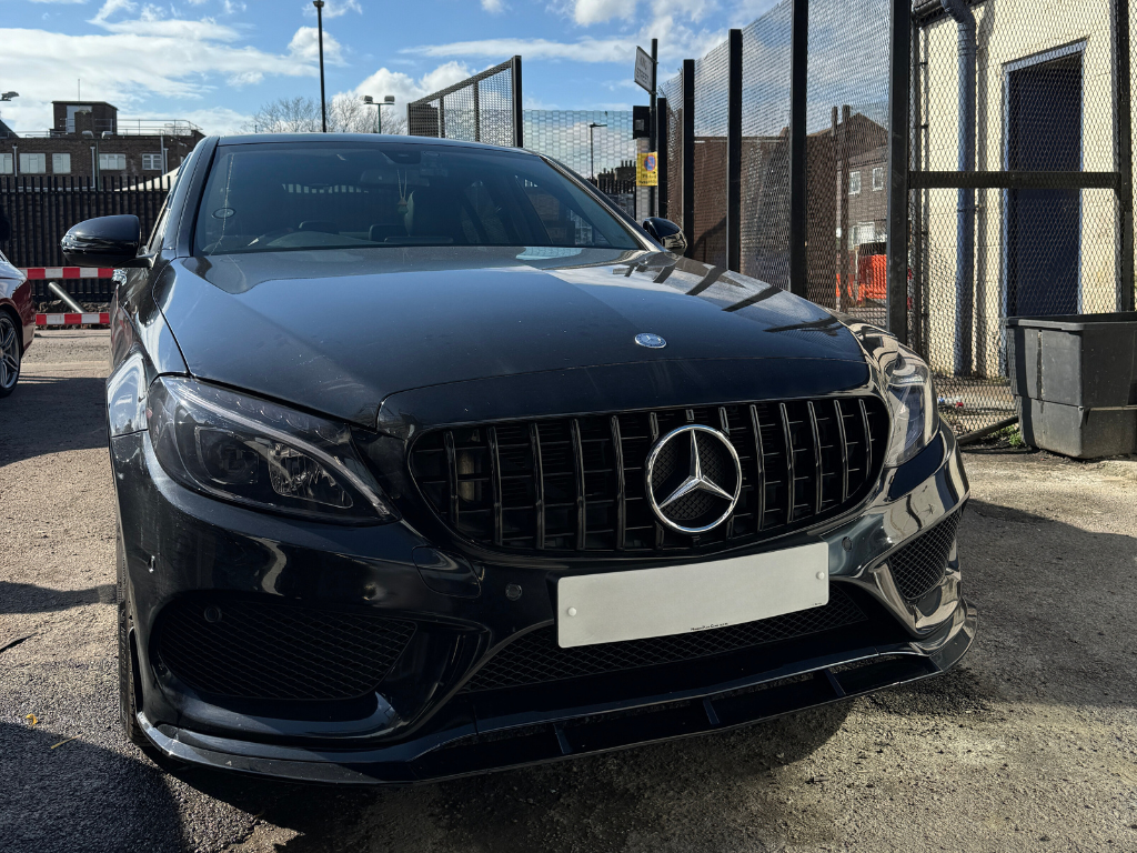 Car parked on asphalt, facing forward with a prominent front grille and emblem. Wire fencing and buildings are in the background under a partly cloudy sky.