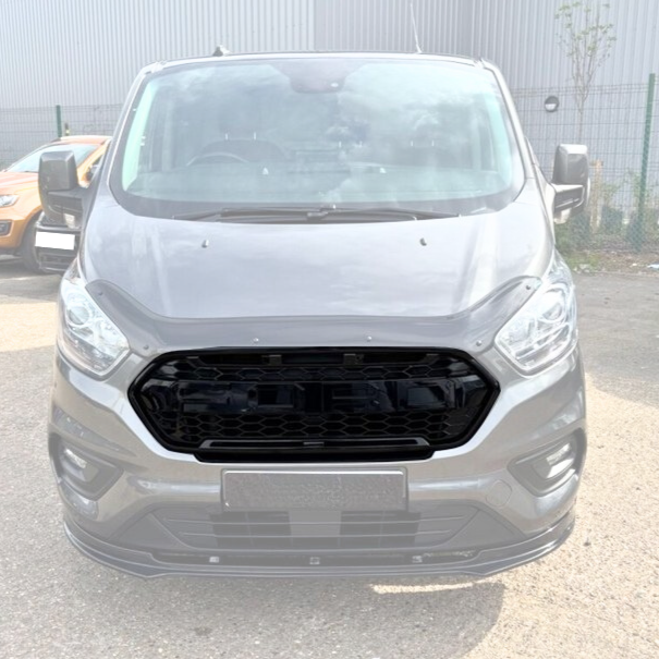 A gray van is parked on a concrete surface, positioned in front of a fenced area and industrial building, with another vehicle partially visible on the left. No text is present.