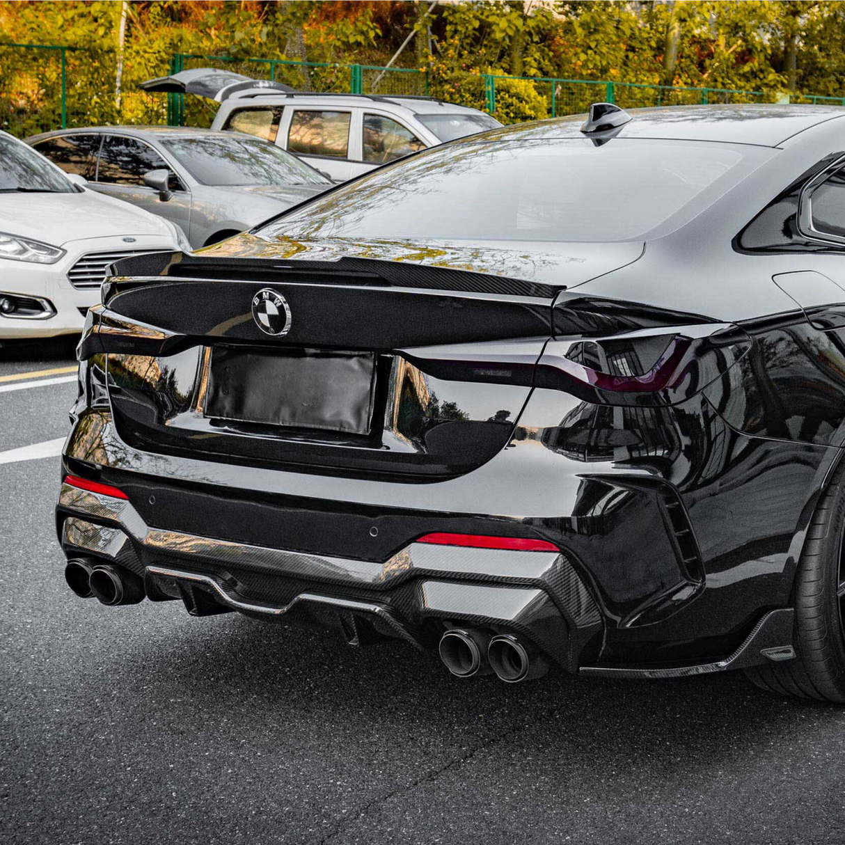 A sleek black car is parked, showcasing its rear with dual exhausts and a BMW logo, surrounded by other parked vehicles and greenery.