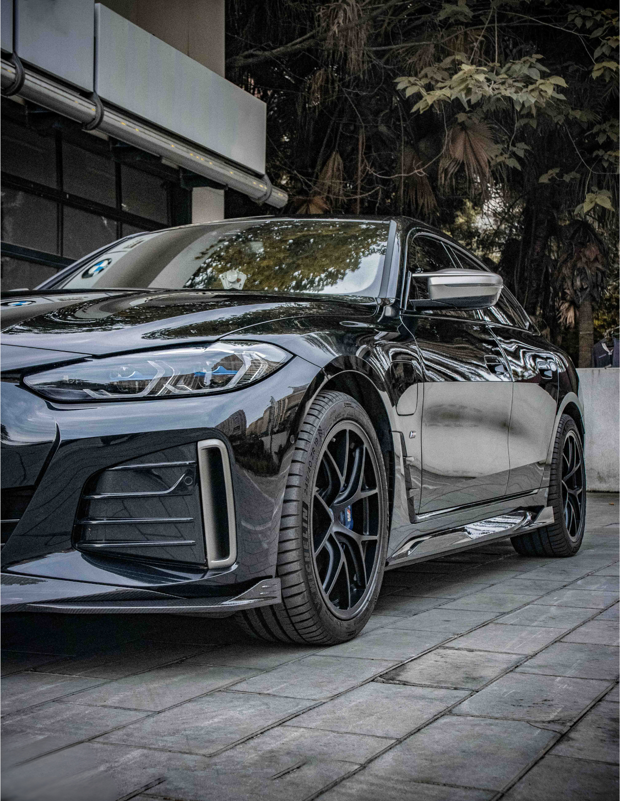 A sleek, black car is parked on a tiled pavement. The setting includes a modern building on the left and lush trees in the background, creating an urban yet natural ambiance.