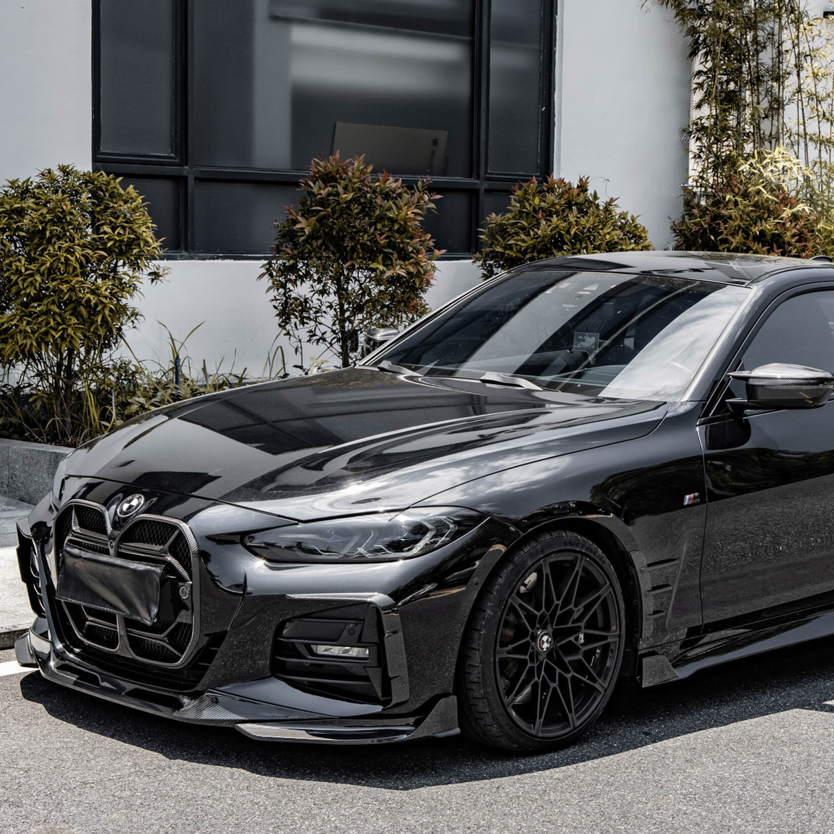 A black car with sleek, sporty design is parked beside a building. Shrubs and greenery provide a contrasting background, highlighting the vehicle's glossy finish and modern lines.