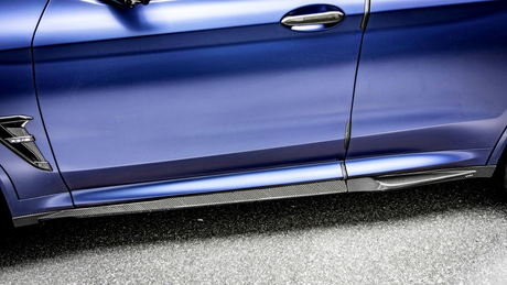 A blue car door with a carbon fiber handle and trim features an "X3 M" badge, parked on a textured gray asphalt surface.