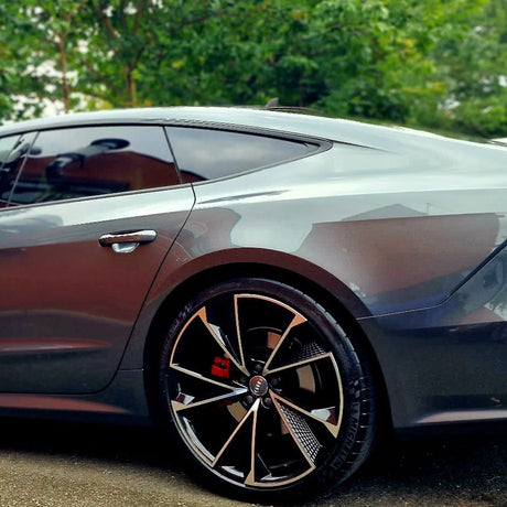 A sleek, metallic gray car, parked with its back wheel in focus, features stylish black alloy rims. Surrounding it are green trees, suggesting a serene, natural environment.