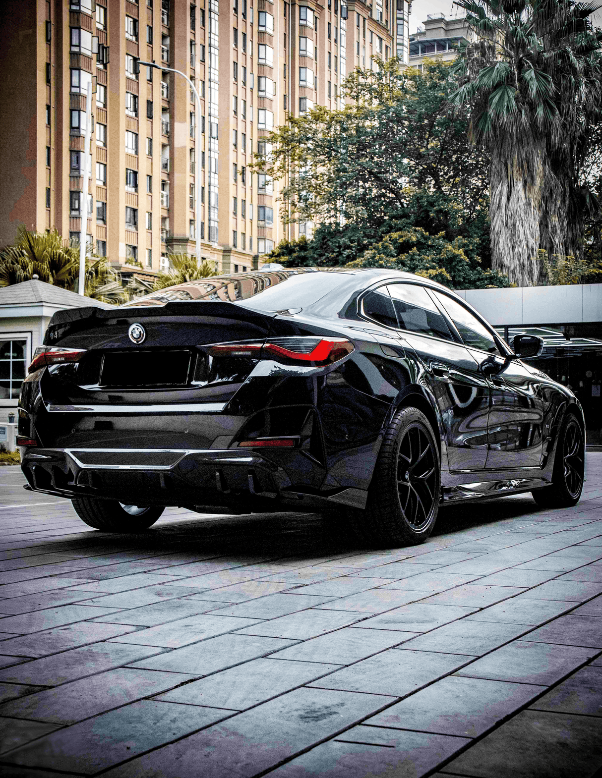 A sleek black car is parked on a tiled sidewalk. Tall buildings and lush trees surround it, creating an urban setting. The detailed design of the car is prominent.
