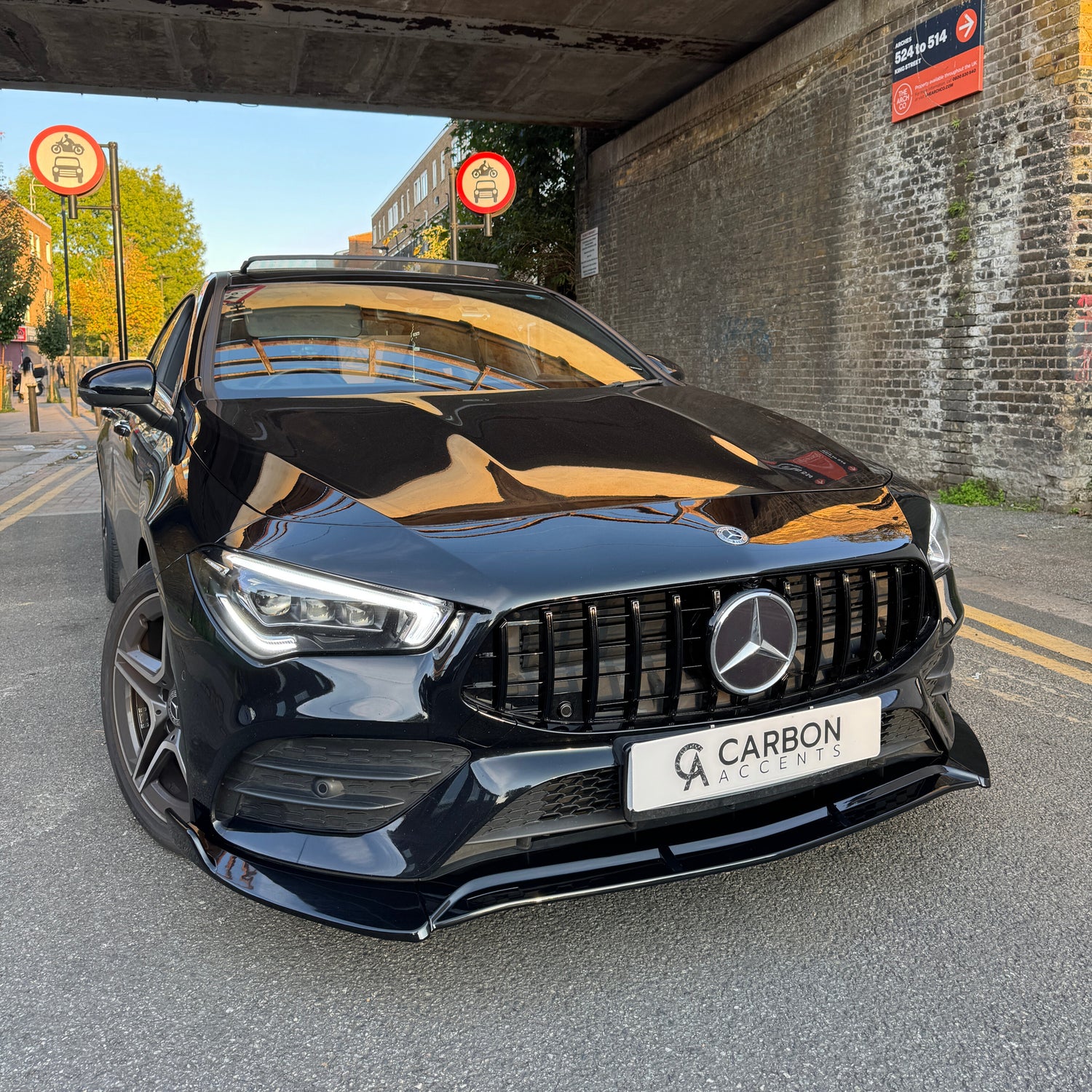 Car: A black Mercedes-Benz, stationary, with a "CARBON ACCENTS" license plate. Context: Parked under a bridge with brick walls, surrounded by "No Cycling" signs and a nearby street with trees and buildings.