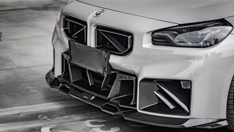 A white car with a sporty front bumper featuring intricate carbon fiber accents, parked in a concrete garage. A covered license plate is visible in the center.