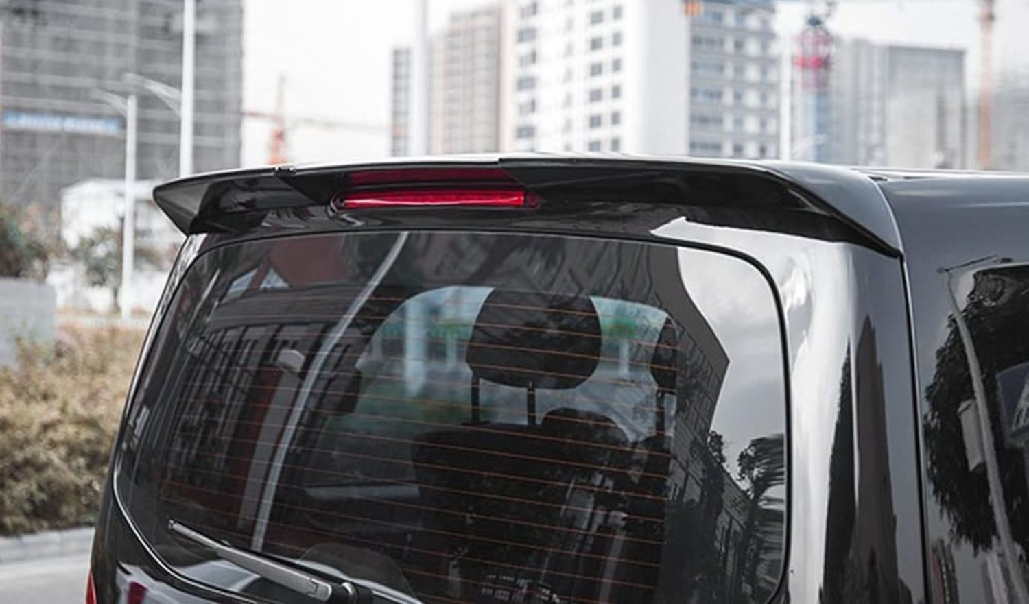 A black vehicle's rear window reflects surrounding trees and buildings, with a red brake light on top. In the background, urban buildings and construction cranes are visible.