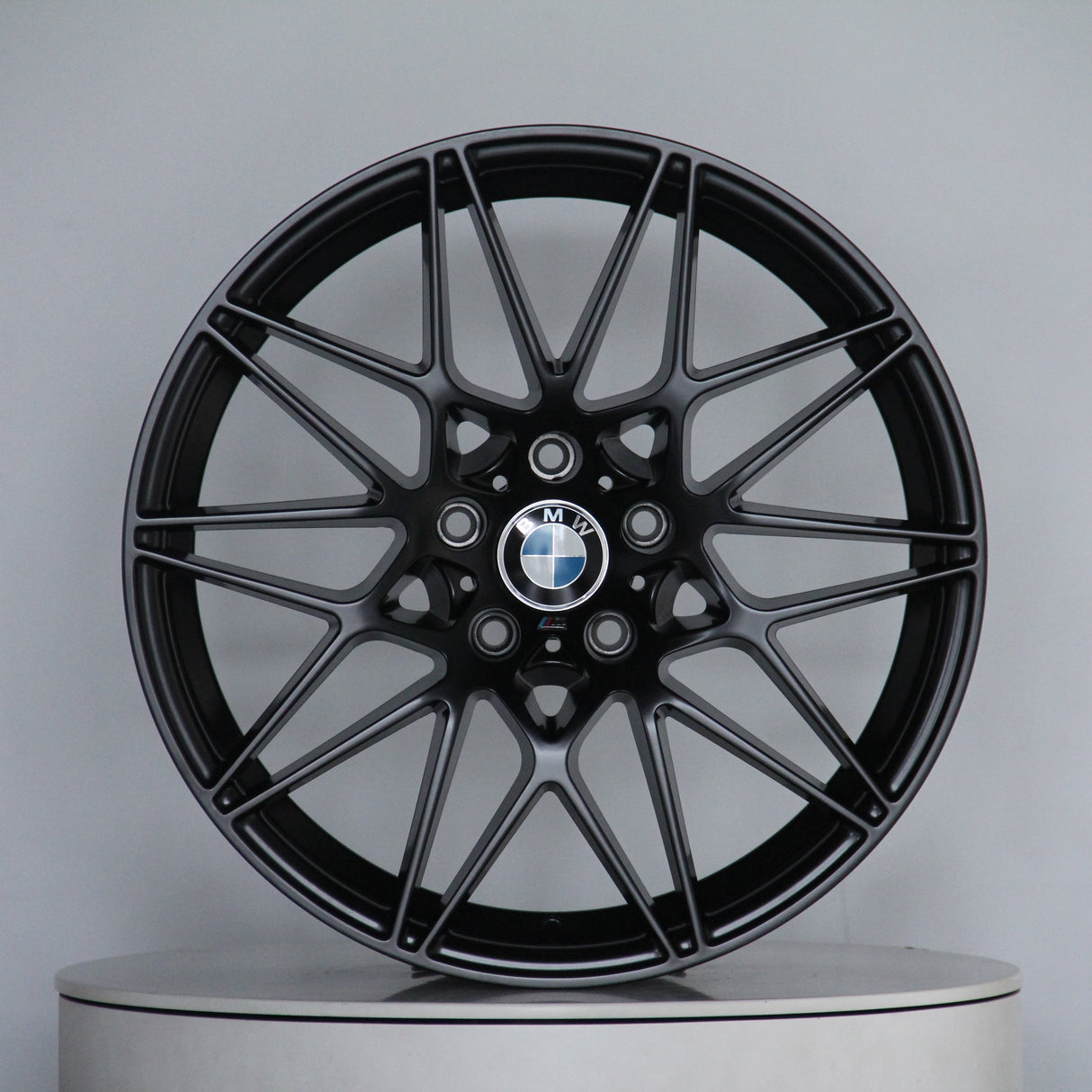 A sleek black alloy wheel with a multi-spoke design, featuring the BMW logo at its center, stands on a plain circular platform in a light gray setting.