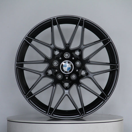 A sleek black alloy wheel with a multi-spoke design, featuring the BMW logo at its center, stands on a plain circular platform in a light gray setting.