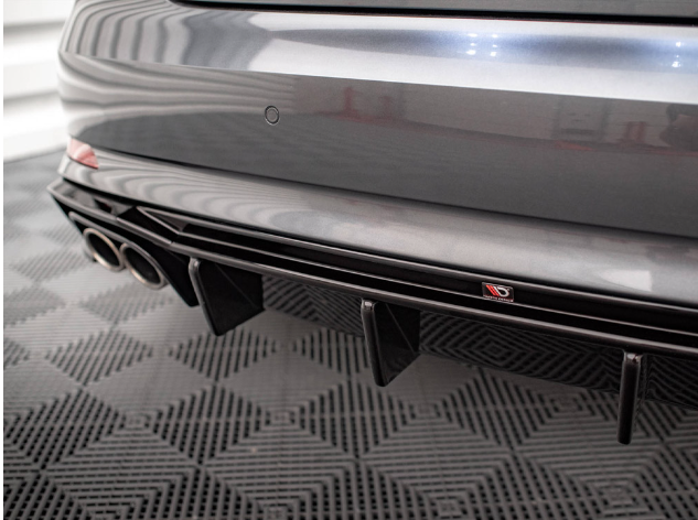 A car's rear bumper with dual exhaust pipes and a diffuser. The car is parked on a textured, black floor, and there's a small emblem displaying a red “D” and “GB”.