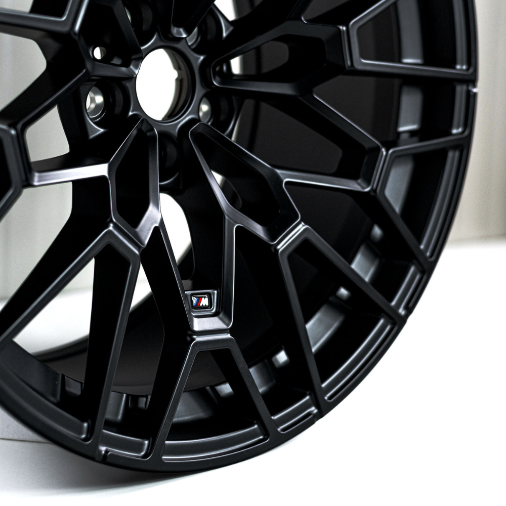 Black car alloy wheel with intricate spokes is displayed against a neutral background. A small emblem with an "M" on it is visible in the center section.