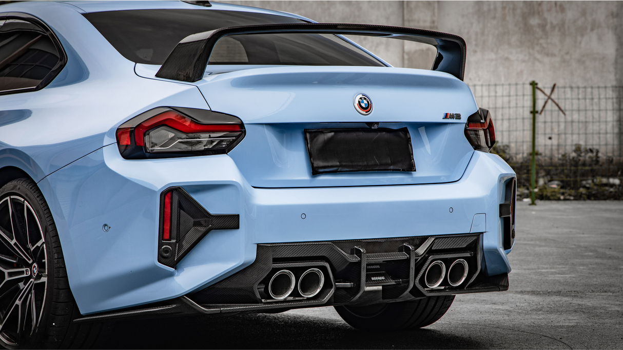 A blue car with a rear spoiler, four exhaust pipes, and a license plate obscured, parked on concrete with a metal fence and wall in the background.