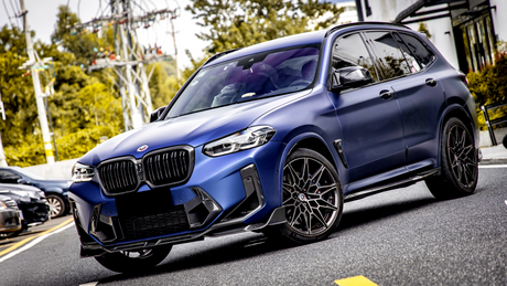 An SUV with a matte blue finish is parked on a street, featuring black grille and wheels, surrounded by trees and parked cars.