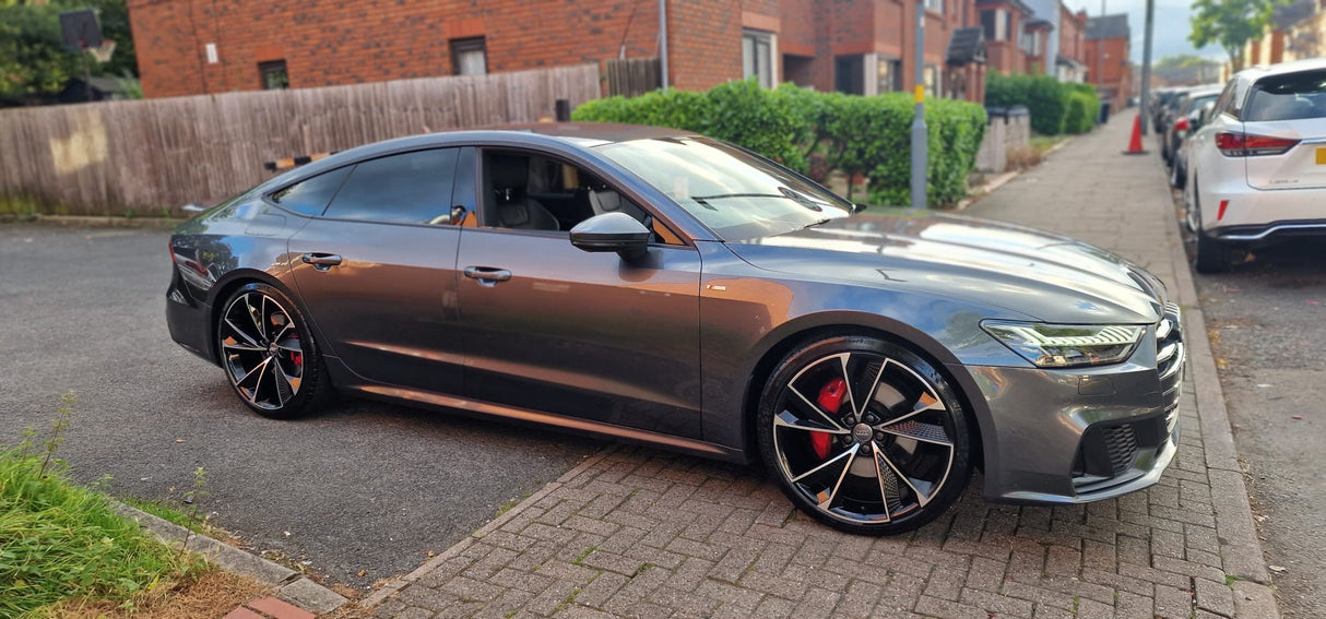A sleek, metallic gray sedan is parked on a residential street, showcasing its modern design and large alloy wheels. Brick houses and a wooden fence line the background.