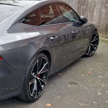 A grey car with black alloy wheels is parked on a paved surface beside a wooden fence, reflecting nearby buildings in its windows.