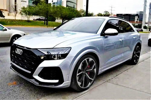 A silver SUV is parked on a street with tall buildings and trees in the background. It's a modern design with large wheels, black accents, and clear glass windows.