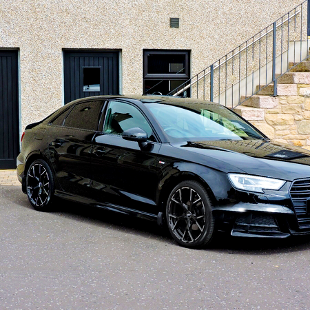 A black sedan parked on a paved surface, with sleek, shiny wheels. It is positioned in front of a stone-textured building with steps and two closed black doors.