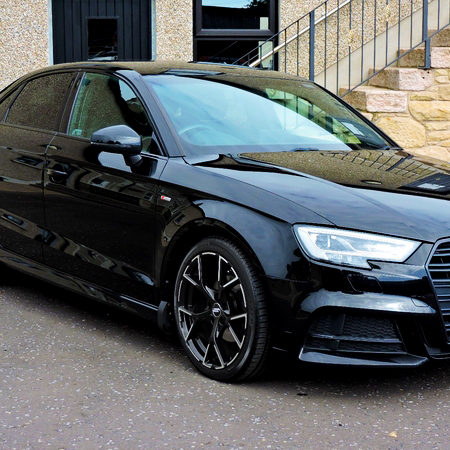 A black car is parked on a paved area near a building with stone and gravel walls, adjacent to metal-railed stairs.
