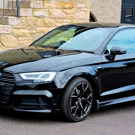 A black car is parked on a street. It is positioned in front of a textured stone and pebble wall, with windows reflecting the surroundings.