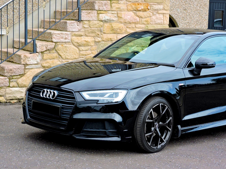A black Audi car is parked on a paved surface. Its sleek design is highlighted, positioned in front of a stone building with exterior stairs.