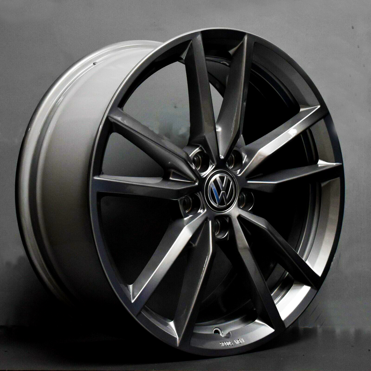 A dark metallic alloy wheel with a Volkswagen emblem rests on a plain surface. Bold, angular spokes create a sleek design. The setting is a simple gray backdrop.