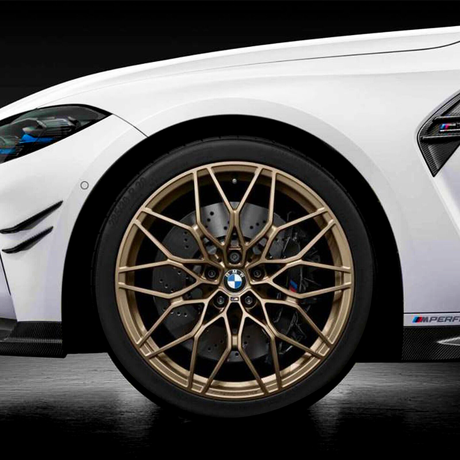 A gold alloy wheel on a white BMW features a complex spoke design. The car is parked in a dimly lit environment with visible "M Performance" badging on the side.