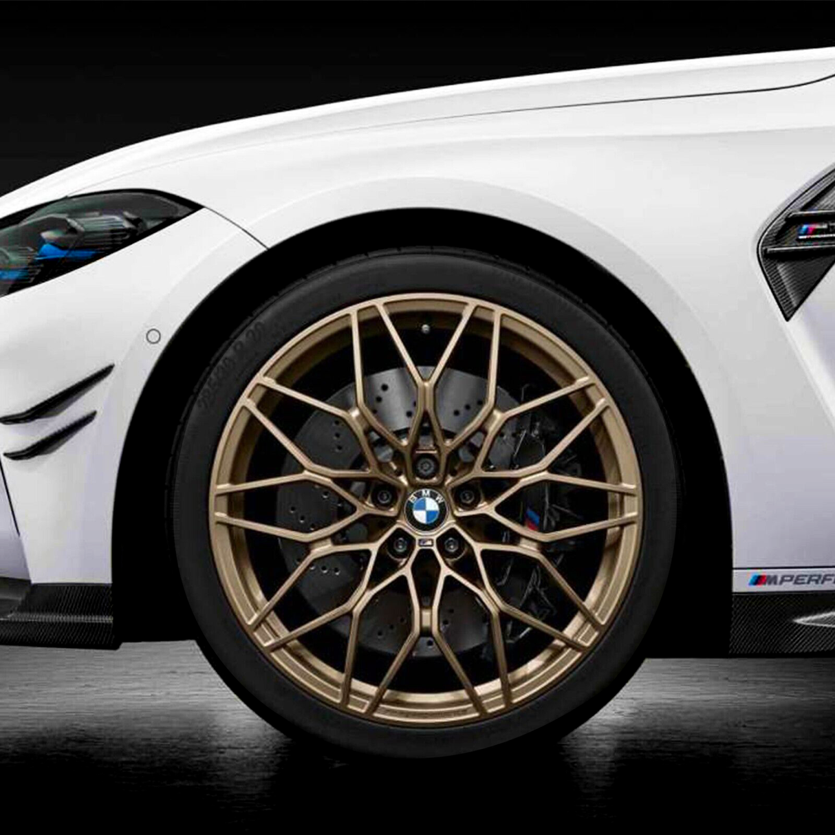 A close-up of a white car's front wheel featuring intricate gold alloy rims with a visible BMW logo, set against a dark background. Carbon fiber accents and "M PERFORMANCE" text are present.