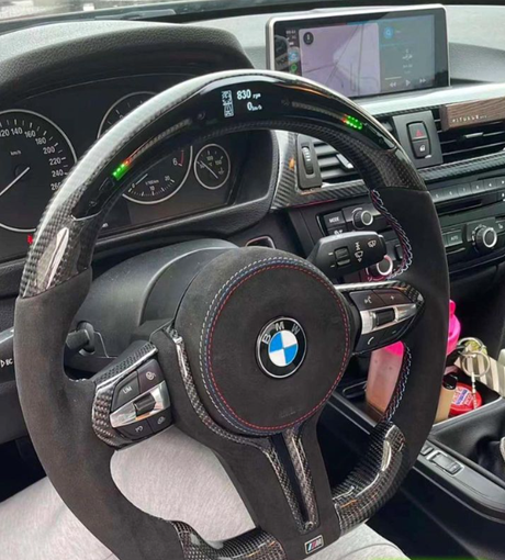 A BMW steering wheel with red stitching displays 830 rpm digitally. Surrounding dashboard shows gauges and controls. The car has a navigation screen and interior accessories on the center console.