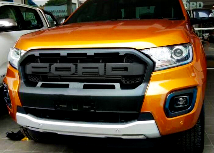 A bright orange truck faces forward, displaying a large black grille with "FORD" lettering. It is parked indoors on a tiled floor, surrounded by other vehicles.
