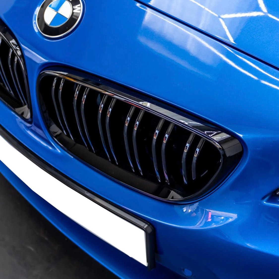 The blue BMW car front shows a kidney grille with vertical slats, featuring the BMW emblem above it, situated in an indoor environment with overhead lighting.