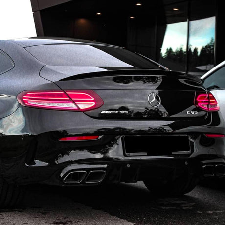 A black Mercedes-Benz AMG C63 parked, with illuminated rear lights, chrome quad exhausts, and badges. It is set against a modern building with reflective windows and dim lighting.