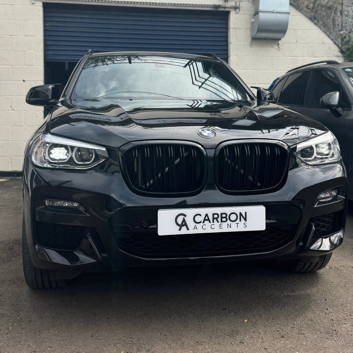 A black BMW car is parked, facing forward. The license plate reads "CARBON ACCENTS." It is situated in a lot with a blue garage door in the background.