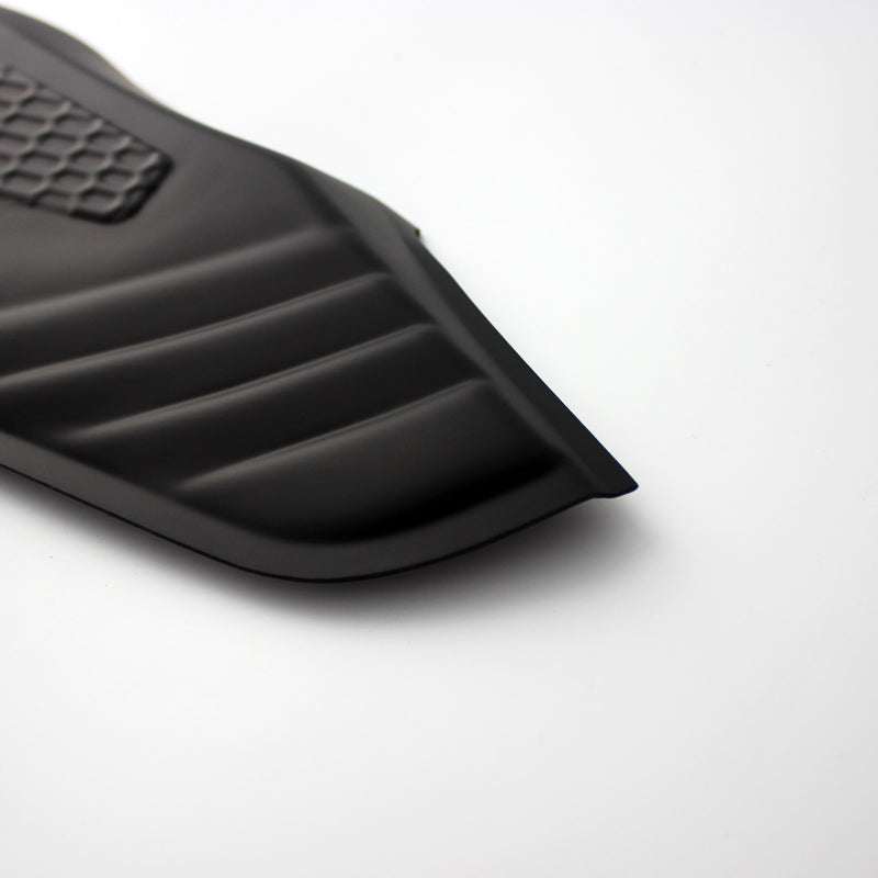 A black, ribbed car floor mat corner with a textured rectangular section rests on a smooth white surface, showing its contoured edges.