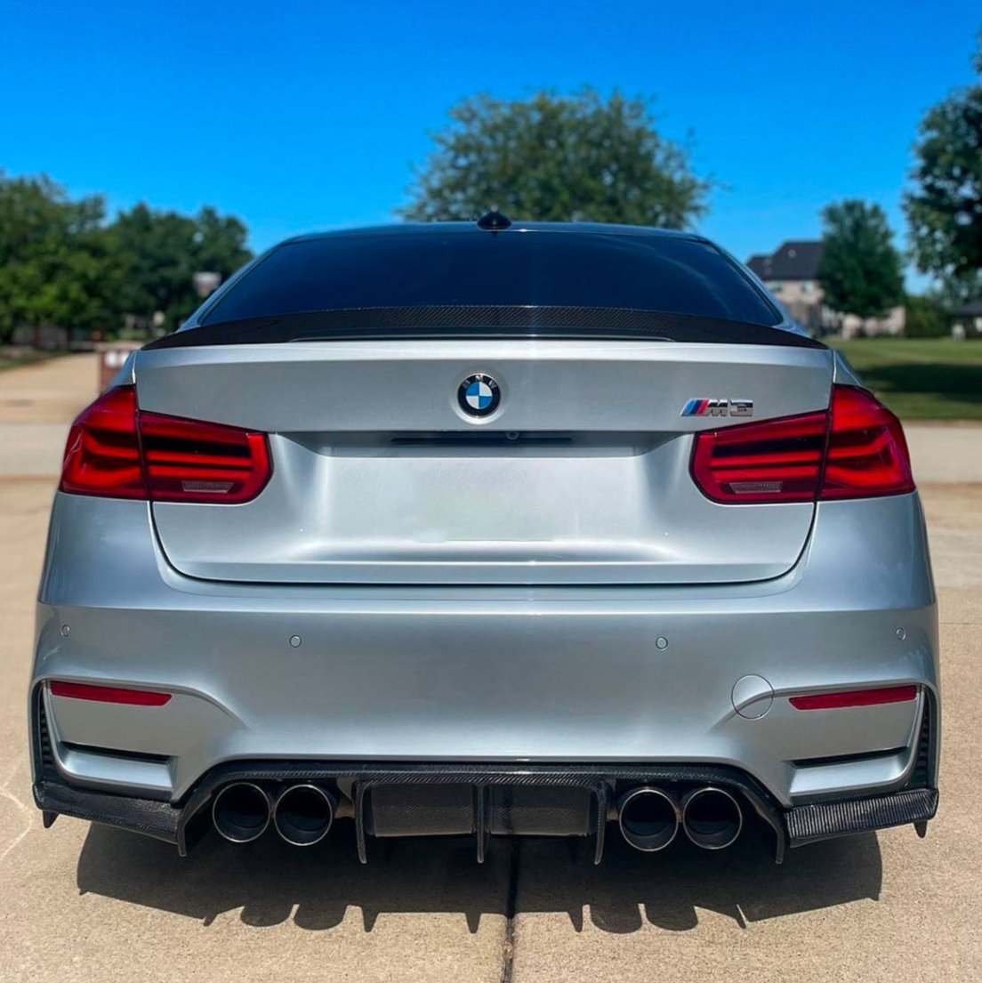 A silver BMW M3 with quad exhaust tips is parked on a driveway. The background features suburban homes and trees under a clear blue sky.