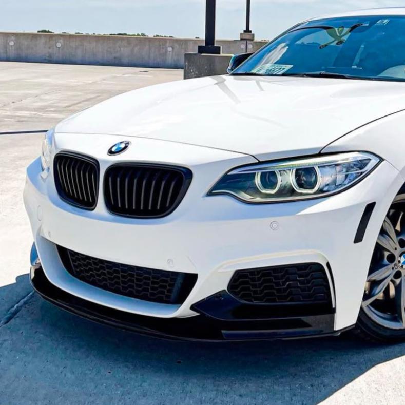 A white BMW car is parked, showcasing its front grille and headlights, in a sunny, open parking lot with a concrete barrier in the background.