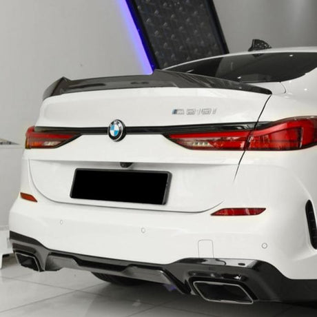 A white BMW 218i is parked indoors, showcasing its rear design with red taillights, dual exhausts, and carbon fiber accents. The setting is a sleek, modern space with bright lighting.